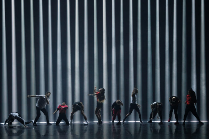 A black stage with a black background and 23 beams of light going straight down, making the back look like prison bars, or a barcode. They are at least 25 feat high, based on the 10 dancers in front of them. The wear black, white and red and are all actively moving. It is as if they are all battling something, from arms punched to the side, above or in front, some are on their knees. 