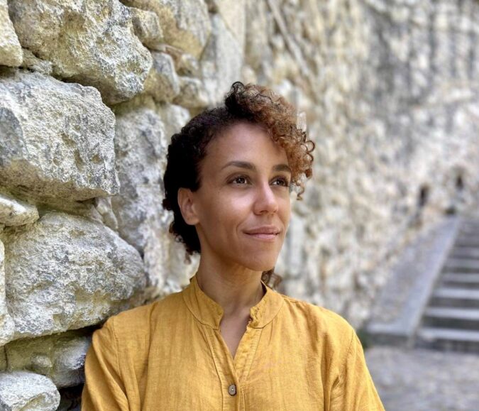 A woman of colour, wearing a yellow blouse stands against a stone wall, looking off camera