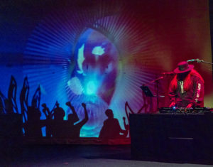 A DJ with long hair wearing a wide-brimmed hat and hoodie performs. The background is a projection of blue, red, and purple abstract shapes with rays beaming out from the centre. Silhouettes of people with raised hands line the bottom.