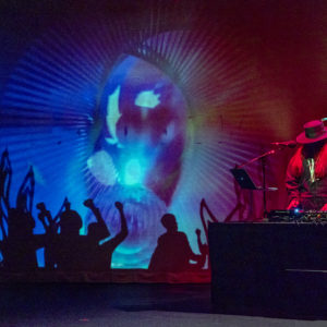A DJ with long hair wearing a wide-brimmed hat and hoodie performs. The background is a projection of blue, red, and purple abstract shapes with rays beaming out from the centre. Silhouettes of people with raised hands line the bottom.