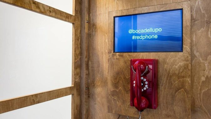 A red telephone with a screen above it that says @bocadellupo #redphone inside of a wooden, handcrafted phone booth