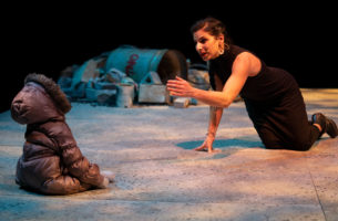 Laara Sadiq, a woman with brown hair in a braid wearing a black blouse, black pants, and black shoes, kneels down and pleads to a mysterious character with the shape of a small child wearing a black parka jacket, gray gloves and gray socks. A pile of gray bricks and a gray bin are placed behind them.