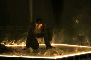 Émilie Monnet, dressed in dark clothing, is onstage on one knee. Wispy smoke emerges from a light-filled pentagram outline on the ground amid a dark room.