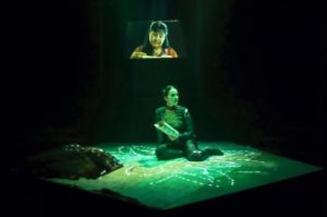 Émilie Monnet, dressed in dark clothing, sits onstage holding a cylinder of bark. On the wall behind is a video of an older woman with bangs holding a similar item. Monet is bathed in green light from above with flower-shaped spots of light projected onto the ground.