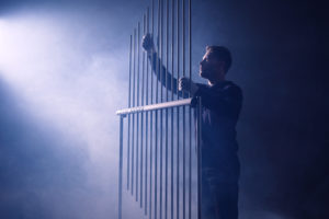 Joby Burgess, with short brown hair and dark clothing, plays a tall aluminum harp. He stands in shadow, with white-purple fog streaming in from the left.