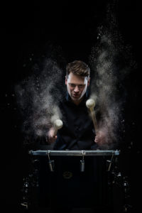 Joby Burgess, who has short brown hair and dark clothing, pounds on a drum with shiny metal hardware using large white-tipped sticks. White dust particles rise dramatically in the air.