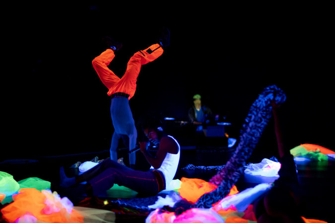 Onstage performance. In the foreground, a performer in a white tank top and holds a microphone; to the left, a dancer in orange pants does a headstand; on the right, a dark-clad figure holds a long piece of fabric. On the ground are neon-coloured tulle costumes. The stage is lit by blacklight, with figures in near darkness and neon colours glowing brightly.