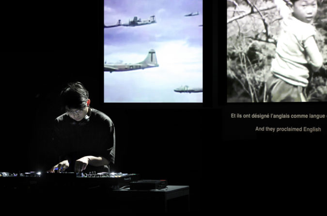 Jaha Koo, who has short black hair and is wearing glasses and a black T-shirt, DJs in a dark room. Behind are two video screens; the left depicts several plans in the sky; the right shows a black-and-white photo of a boy with the text “Et ils ont désigné l’anglais comme langue / And they proclaimed English”.