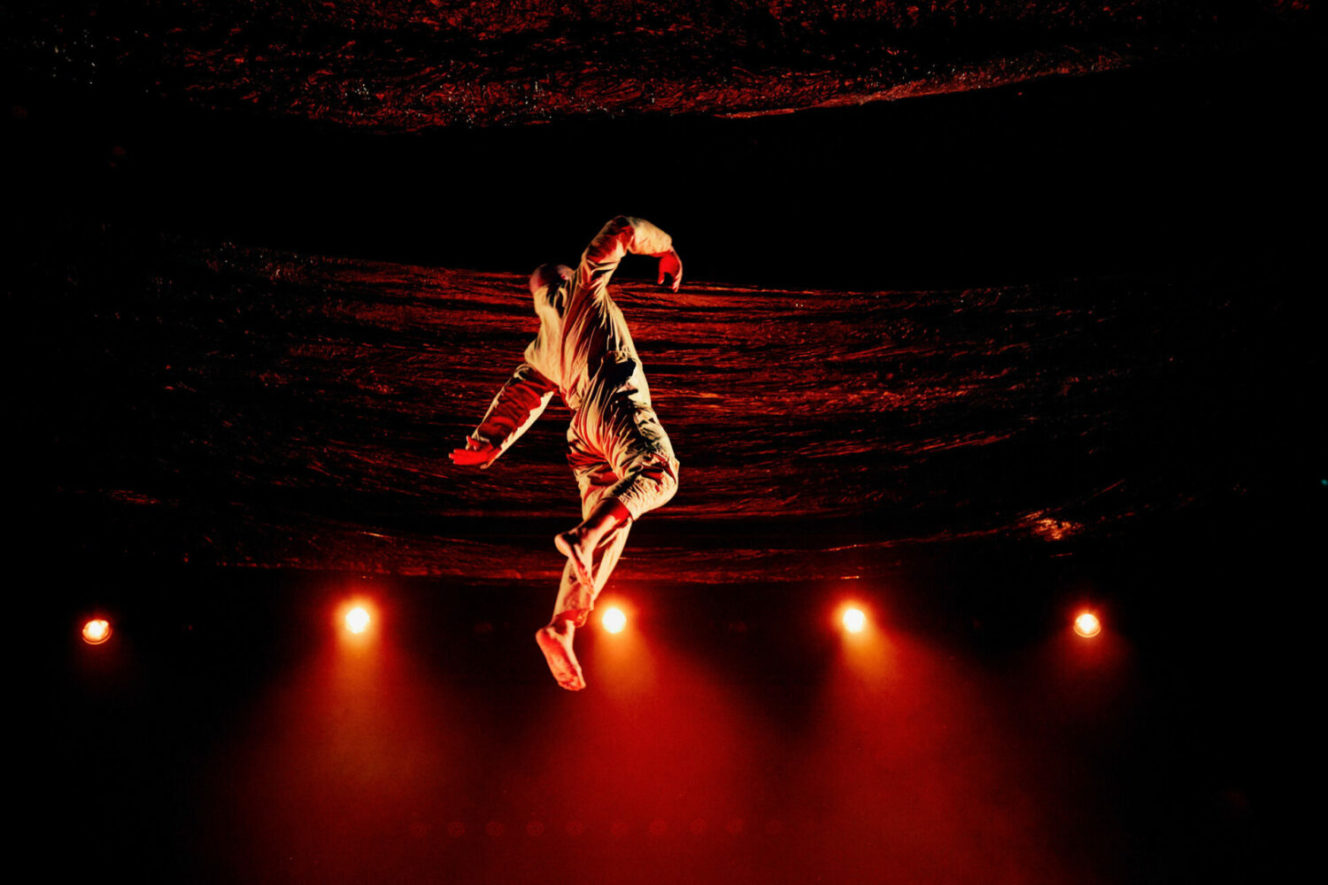 A performer, barefoot and wearing a yellow jumpsuit, jumps high in the air with arms gesticulating. In the background are red stage lights and red fabric hangings.