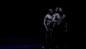 Three men, shirtless with words painted on their bodies, stand on a black background making gestures