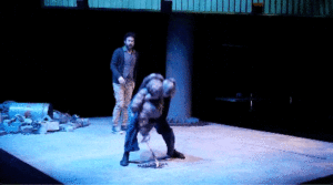 A woman picks up a misshapen, human-like infant form and a man watching turns and runs away. The scene changes to a small group of people standing in a circle on a dimly lit stage, arms raised.
