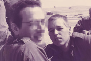 Duotone purple and beige photograph of two male figures, both with short hair and wearing military uniforms. Both are looking towards the camera, but the face in the foreground is pixelated. In the background are desert hills and partial views of other figures.