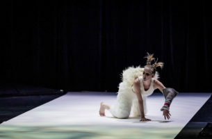A performer with blonde pigtails wears dark eye makeup, white tank top, furry white paints, and a grey knitted sleeve on the left arm. They are kneeling on the ground looking up. The ground is lit with white lights amid a dark stage.