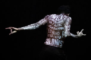 A dancer in black pants stands facing away from the camera, arms gesturing. The room is darkly lit, highlighting their bare back and arms which are covered in white-painted testimonials of the families of young Black men who have been victims of gun violence.