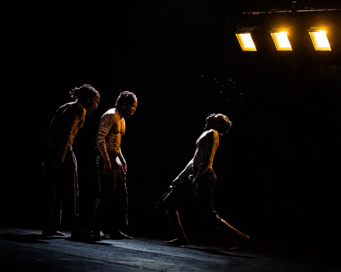 Three shirtless dancers wearing black pants stand facing each other onstage, with bare torsos covered in white-painted testimonials of the families of young Black men who have been victims of gun violence. Two figures on the left face the one on the right, who is lunging and looking up with eyes closed. Beads of sweat are visible under the stage lights.