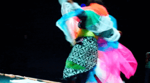 A person dressed in many overlapping colourful textiles dances in circles
