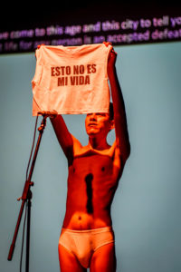 Tiziano Cruz stands in front of a microphone, wearing only white briefs, holding up a tank top with the words “ESTO NO ES MI VIDA” on it. In the background is a grey screen with out-of-focus purple text.