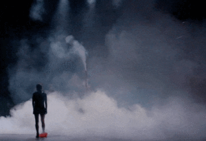 Short video clip of a person walking on stage into a smoke filled background while another person blows smoke from a hose.