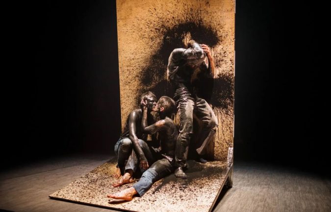 Two dancers covered in black paint sit shirtless, kissing, on top of a freestanding wood structure. A third, also covered in black paint, stands above them with their head in their hands, leaning against a paint-splattered wood wall.