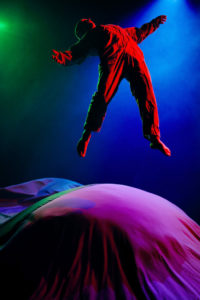 Dramatically lit image of a performer, barefoot and wearing a jumpsuit, jumping midair and against a blue-lit background. The figure is bathed in red light and below are large round fabric structures in purple, green, and blue.