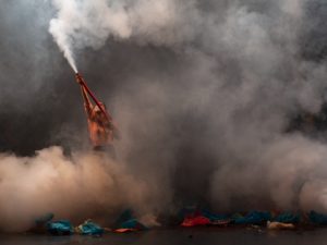 A figure wearing a salmon-coloured shirt and face mask holds up a red tube exhuming a large cloud of white smoke that envelops the room. On the ground are multicoloured pieces of crinkled plastic.