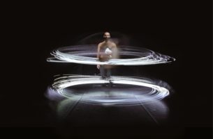 Long-exposure image of Juan Ignacio Tula in Instante, standing in a dark room. He is shirtless and stares into the camera, holding a white stack of paper. Circling him are two rings made of many horizontal rings of light motion lines.