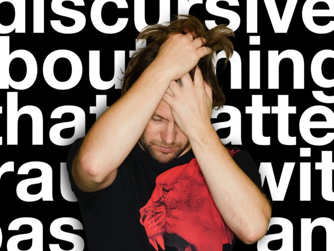 Joby Burgess, wearing a black shirt with a red roaring tiger on it, runs his hands through his brown hair. Behind is a black background with oversize white words that are not legible.