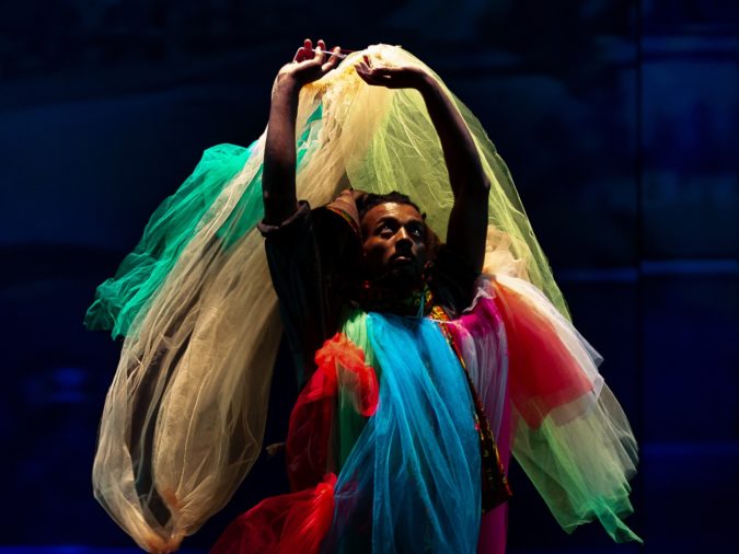 A dancer wearing a costume of brightly coloured flowing tulle stands with arms raised, looking up. They are lit by light above amid a dark blue background.