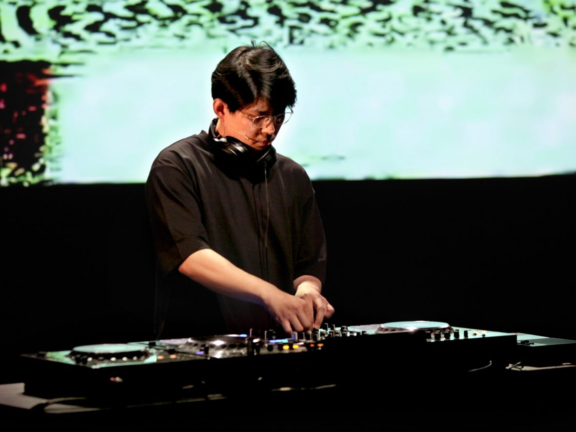 Jaha Koo, who has short black hair and is wearing glasses and a black T-shirt and headphones around his beck, is DJing. In the background is a video of green glitches.