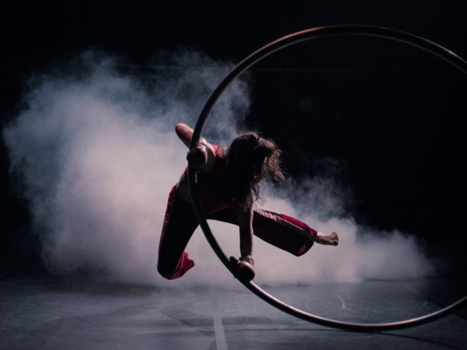 Lontano performance: Marica Marinoni, who has wavy dark hair and is wearing red pants, is suspended in the air. She holds onto a cyr wheel spinning against the ground amid a room filled with fog.