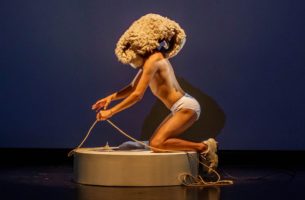 Performer Tiziano Cruz, wearing white briefs and white sneakers, kneels on a small round platform. He pulls on a piece of rope and is wearing a large woolen piece on his head, obscuring the face.