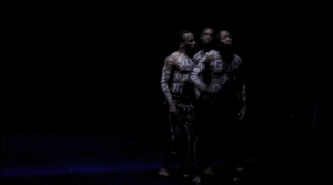 Three men, shirtless with words painted on their bodies, stand on a black background making gestures