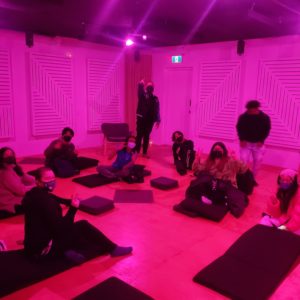 A group of youth sit in a room bathed in red light. The group is sitting on pads on the floor in a semi-circle, facing the camera. Two people are standing and one makes a peace sign to the camera.