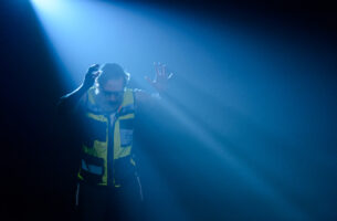 A man in a reflective safety jacket with his hands up and head tilted down. There is a light casting down on him creating light shadows in the image.