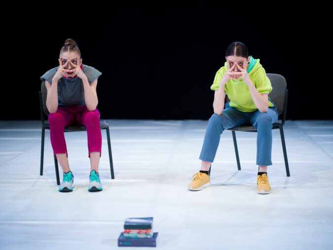 Two performers holding their faces to show their wide eyes. They are both sitting on chairs with their feet apart. There is a stack of books in the foreground of the image.