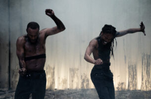 Two Black performers captured intensely mid-dance. The Black man on the left has a buzzcut, one of his arms is raised, with both hands in a fist. The Black man on the right has long hair, posing similarly, except with one hand open. They are both looking at the ground.