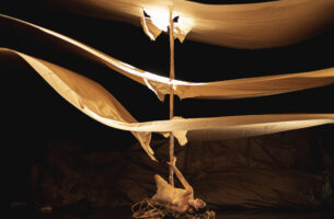 A performer lies on a pile of twisted branches holding up a wooden pole struck through three floating layers of paper.
