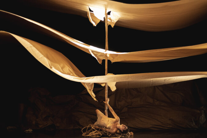 A performer lies on a pile of twisted branches holding up a wooden pole struck through three floating layers of paper.