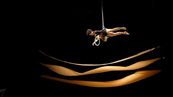 A person with dark curly hair dressed in black is stiffly suspended in the air, harnessed by and holding thick rope. Below are three undulating swaths of paper suspended horizontally.