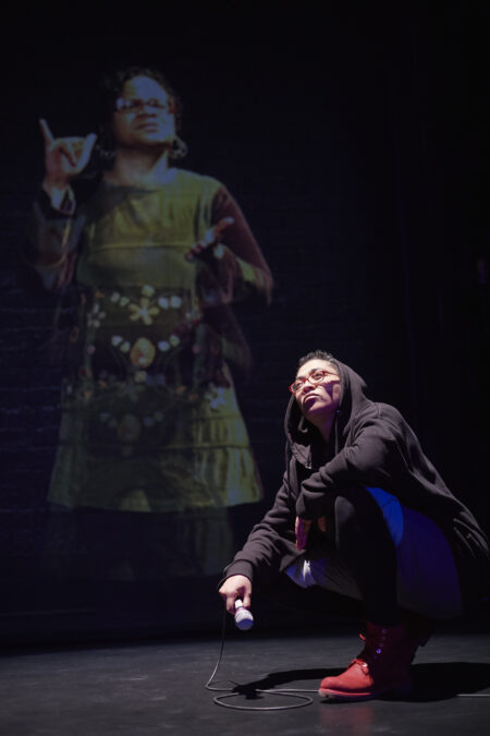 A person half-kneeling with crossed arms while holding a mic. The person is looking up at the light, There is a woman projected behind them.