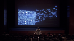 A wideshot of a person facing a projection on a stage. The projection is a glowing blue square pattern. There are light blue shards floating out from the square. The person is standing in the front row, with their arms slightly lifted on their sides.