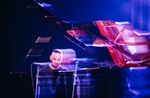 A photo of a man playing the piano. The photo is slightly blurred, the man faces the camera with what appears to be a melancholic expression.