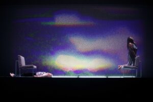 Two people onstage. On the left a person with white beard lies down under a grey couch. On the right, a person with long braided hair kneels on a similar couch facing away, looking to the sky. The backdrop is a grainy, abstract image of purple and white ambient light.