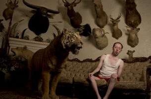 A man sits on a couch leaning away from what appears to be a taxidermy tiger. Behind him is a wall of mounted animal taxidermy heads.