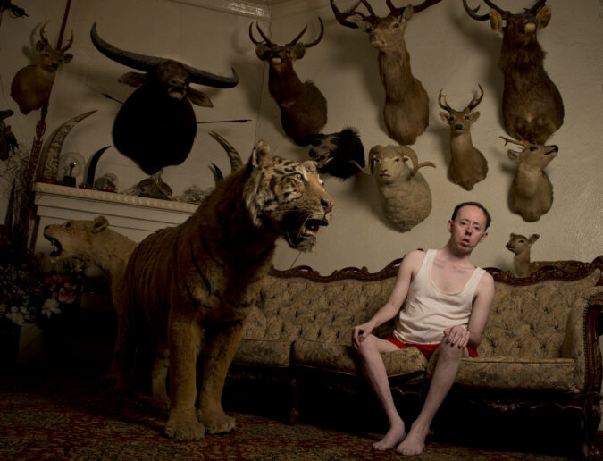 A man sits on a couch leaning away from what appears to be a taxidermy tiger. Behind him is a wall of mounted animal taxidermy heads.