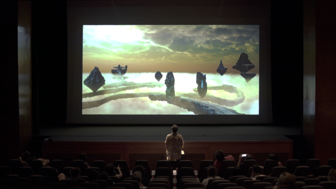 A person stands up in a theatre facing the screen, with seated audience members around them. On the screen are silhouetted floating islands atop a layer of clouds, with sunlight fiercely shining through heavy clouds.