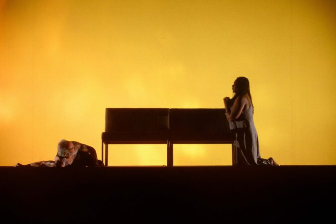 A Black woman kneels against a couch in a praying stance before a yellow backdrop. An older man with grey beard lays on the ground below, arm outstretched.