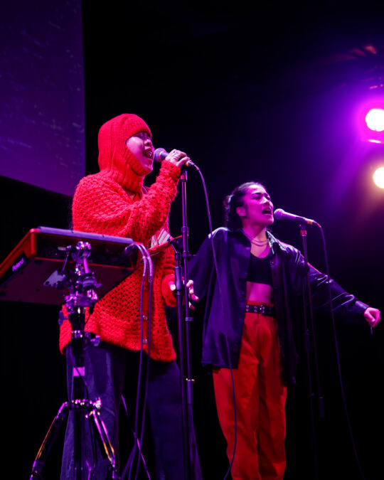 Artists Kimmortal and Ariane Custodio singing onstage, bathed in pink and red light.