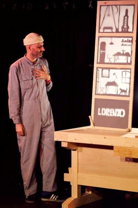 Ben Target, wearing white beanie and boiler suit, holds their chest while looking down at a wooden desk. On the desk sits a wooden panel with silhouetted paper-cut artwork.