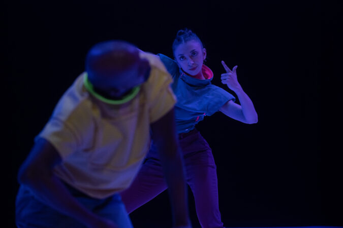 Wide shot of two performers dancing in the dark. In the foreground, out of focus, the performer is hunched over. In the background, the performer looks directly into the camera, hip cocked while making a finger-gun pose.